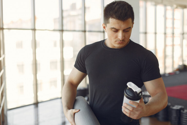 Homem analisando shaker na academia, refletindo sobre os benefícios da bebida proteica pronta para incluir na rotina de treinos.