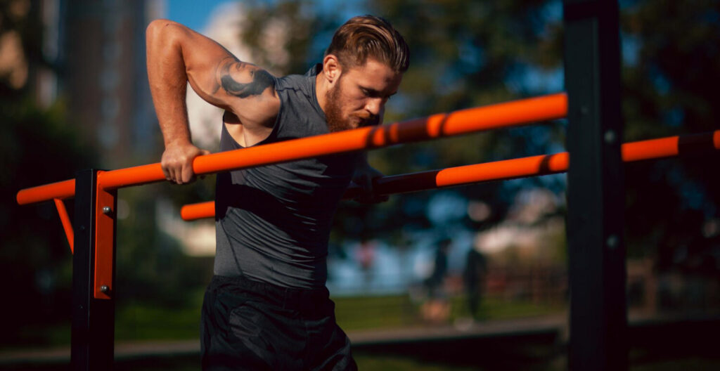 Homem realizando exercício em barras paralelas ao ar livre, treino de força que contribui para o desenvolvimento da resistência física e aumento da performance muscular.