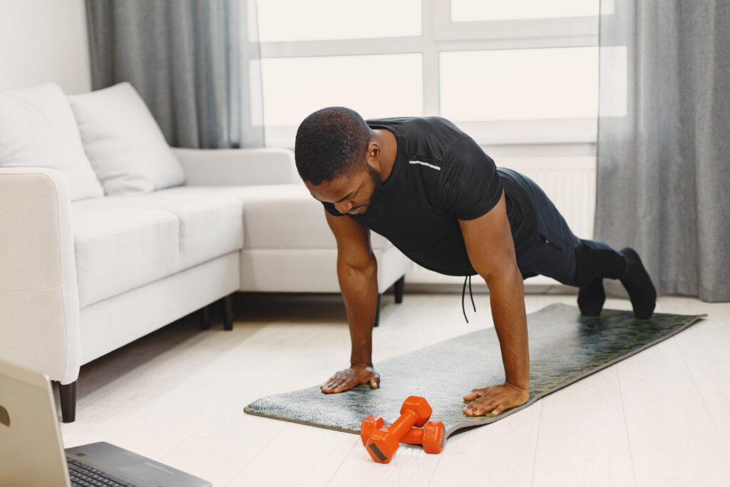 Homem fazendo flexão de braços durante treino em casa, utilizando tapete e acompanhando exercício por notebook.
