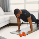 Homem fazendo flexão de braços durante treino em casa, utilizando tapete e acompanhando exercício por notebook.